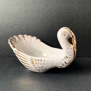 May include: White ceramic swan-shaped dish with gold accents. The dish features a detailed design with gold lines and dots on the body and wings. The swan's head is upright, and the dish is on a dark surface.