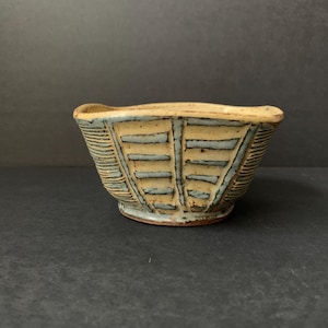 May include: A small, handmade ceramic bowl with a unique design. The bowl features a beige base color with blue rectangular and line patterns. The rim has a slightly irregular shape. The bowl is likely used for serving or display.