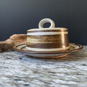 May include: A ceramic butter dish with a lid and a matching plate. The dish is cylindrical with a handle on top, and the lid and plate are decorated with horizontal brown stripes on a cream background. The dish is on a marble surface.