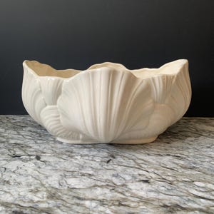 May include: A cream-colored ceramic bowl shaped like a large seashell. The exterior features a repeating embossed scallop shell pattern with a glossy finish and a wavy, scalloped rim. The bowl has a wide, open interior and a smooth, curved base.