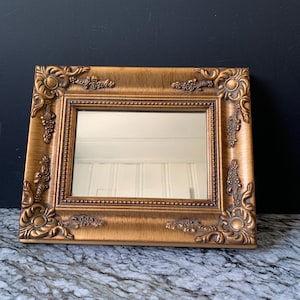 May include: A rectangular mirror featuring a wide, ornate gold-colored frame with intricate embossed scrollwork and floral detailing at each corner. The inner edge of the frame is lined with a delicate beaded texture surrounding the reflective glass surface.