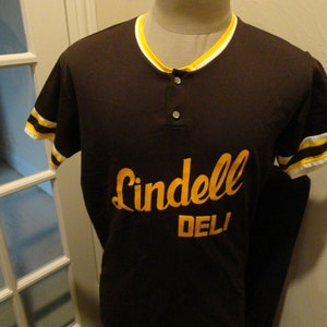 Può includere: Una maglia da baseball marrone con strisce gialle e bianche sulle maniche. La maglia ha un colletto abbottonato e dice "Lindell Deli" in caratteri gialli.
