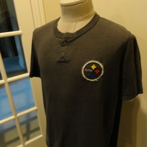 Può includere: Una maglietta a maniche corte di colore grigio scuro con un logo nero, giallo e bianco dei Pittsburgh Steelers sul davanti. La maglietta ha un colletto abbottonato.