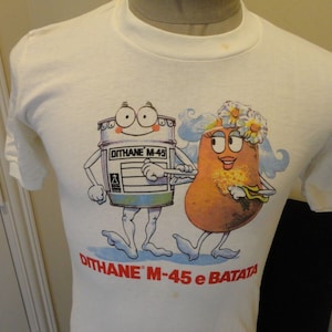 以下が含まれることがあります： Dithane M-45と花冠をかぶったジャガイモの漫画イラストが描かれた白いTシャツ。イラストの下に「Dithane M-45 e Batata」という文字が印刷されています。