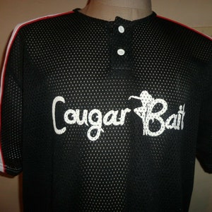 Puede incluir: Jersey de béisbol de malla negra con ribetes blancos y rojos. El jersey tiene un gráfico blanco de una mujer con el texto "Cougar Bait" en la parte delantera.