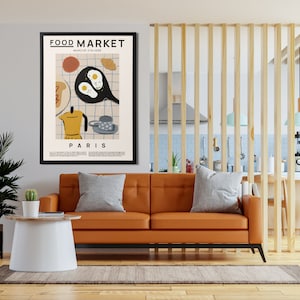 May include: A framed poster with a minimalist illustration of a breakfast scene. The poster is titled "FOOD MARKET" and "PARIS" and features a frying pan with eggs, a coffee pot, a croissant, and a cup of coffee. The poster is hanging on a wall in a living room setting.