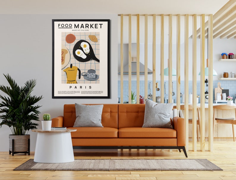 May include: A framed poster with a minimalist illustration of a breakfast scene. The poster is titled "FOOD MARKET" and "PARIS" and features a frying pan with eggs, a coffee pot, a croissant, and a cup of coffee. The poster is hanging on a wall in a living room setting.