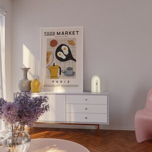 May include: A white dresser with three drawers and gold handles. A white arch lamp with a gold accent sits on top of the dresser. A poster with the text "FOOD MARKET" and "PARIS" hangs on the wall above the dresser. A vase with purple flowers sits on a pink round table in front of the dresser.