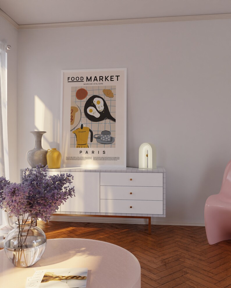 May include: A white dresser with three drawers and gold handles. A white arch lamp with a gold accent sits on top of the dresser. A poster with the text "FOOD MARKET" and "PARIS" hangs on the wall above the dresser. A vase with purple flowers sits on a pink round table in front of the dresser.