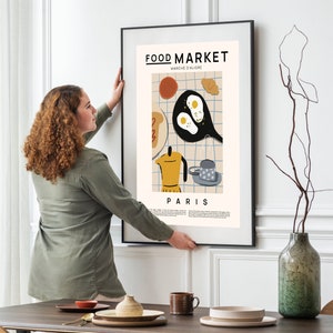 May include: A woman hangs a framed poster on a white wall. The poster features a colorful illustration of breakfast food with the text "FOOD MARKET MARCHE D'ALIGRE PARIS".