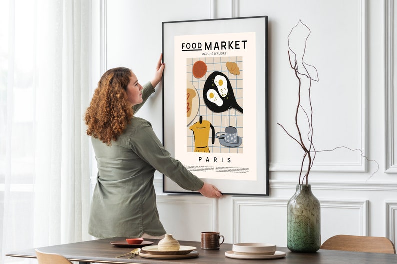 May include: A woman hangs a framed poster on a white wall. The poster features a colorful illustration of breakfast food with the text "FOOD MARKET MARCHE D'ALIGRE PARIS".