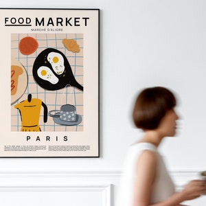 May include: A poster with a black frame featuring a colorful illustration of breakfast food, including eggs, bread, coffee, and a croissant. The poster has the text "FOOD MARKET" and "MARCHE D'ALIGRE" in black text. The poster also has the text "PARIS" in black text.