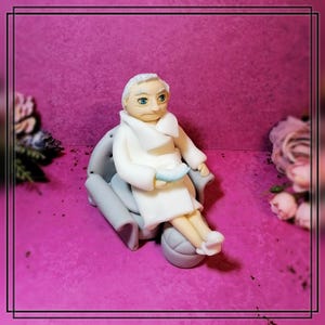 Könnte beinhalten: Eine weiße Fondantfigur einer Person, die in einem grauen Sessel sitzt. Die Person trägt eine weiße Robe und hat weißes Haar. Die Person hält eine kleine weiße Schüssel in ihren Händen.