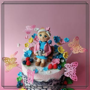 Könnte beinhalten: Ein dekorierter Kuchen mit einer Figur einer Frau, die auf einer Bank sitzt und eine Tasse hält. Der Kuchen ist mit bunten Blumen, Beeren und Schmetterlingsdekorationen verziert. Der Kuchen hat einen rosa und weißen Zuckerguss mit einer dunkelgrauen Basis.