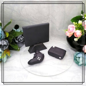 May include: A black and white cake topper featuring a television, a video game console, and a controller. The cake topper is on a clear round cake stand.