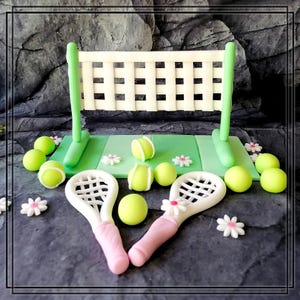 Könnte beinhalten: Ein grüner und weißer Tennis-Kuchentopper mit Netz, zwei Schlägern und mehreren Tennisbällen. Die Schläger sind rosa und weiß, die Bälle sind gelb.