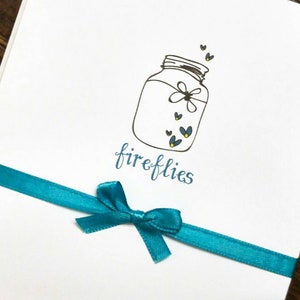 May include: A white card with a blue ribbon tied in a bow. The card has a hand-drawn illustration of a mason jar with fireflies inside. The text "fireflies" is written below the jar.