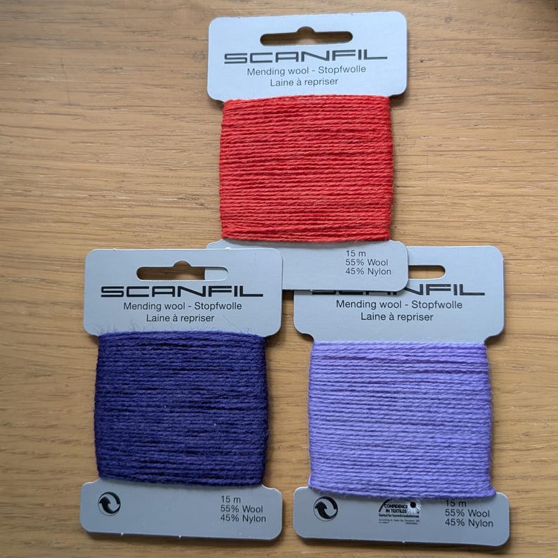 May include: Three Scanfil mending wool cards in red, dark purple, and light purple. Each card contains 15 metres of yarn made from 55% wool and 45% nylon. The white cardboard packaging features the brand name and product details in multiple languages.