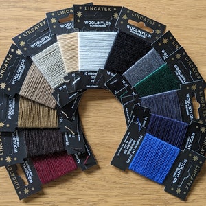 May include: A variety of 10-meter spools of wool/nylon thread for mending, in colors including beige, brown, black, red, blue, green, and gray. The spools are arranged in a semi-circle on a wooden surface. Each spool has a black label with the text "LINCATEX WOOL/NYLON FOR MENDING" and the length of the thread in meters.