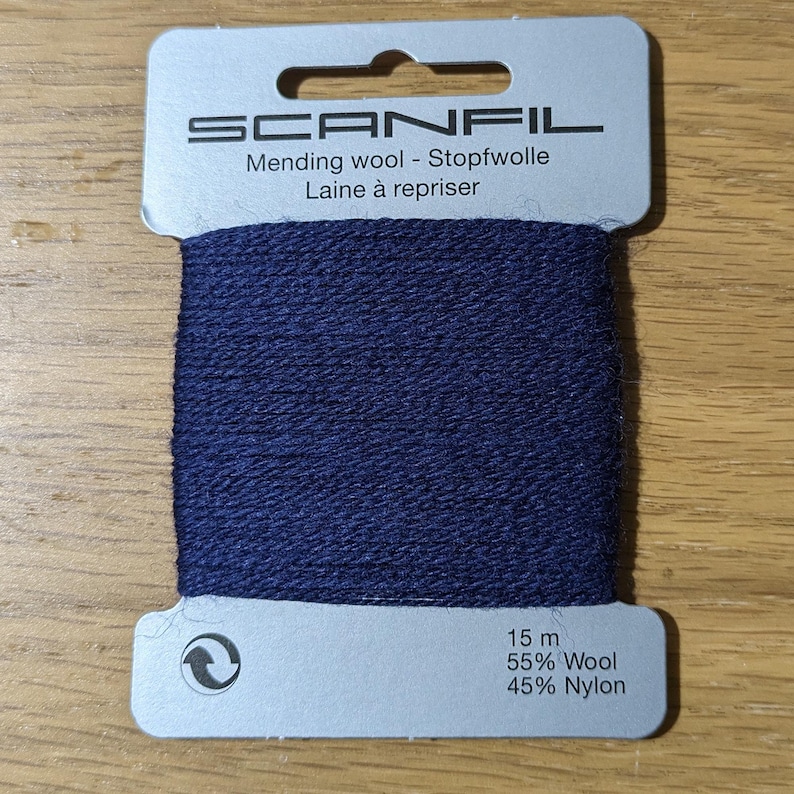 May include: A card of navy blue Scanfil mending wool wrapped around a light grey cardboard holder. The text reads Scanfil, Mending wool, Stopfwolle, Laine &agrave; repriser, 15 m, 55% Wool, and 45% Nylon. The yarn has a textured, matte finish.