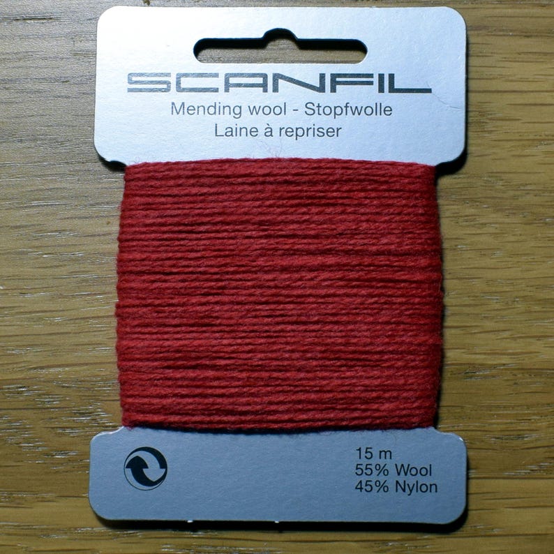 May include: A card of red mending wool by Scanfil. The light grey cardboard holder features the brand name, the text Mending wool - Stopfwolle Laine &agrave; repriser, and technical details stating 15 m, 55% Wool, and 45% Nylon with a recycling symbol.