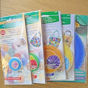 May include: Four packs of Clover brand "Quick" Yo-Yo Makers. Each pack features a different colour scheme and includes instructions. The packs are labelled with the product name, art number, and size of the yo-yos. The packs are on a wooden surface.
