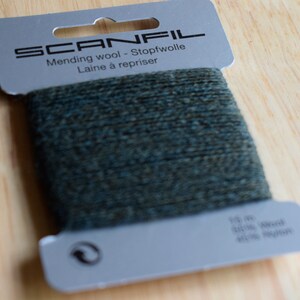 May include: A card of dark green mending wool by Scanfil. The grey cardboard packaging features the text SCANFIL, Mending wool - Stopfwolle, Laine &agrave; repriser, 15 m, 50% Wool, 50% Nylon, and a recycling symbol. The dark green yarn is wrapped around the centre.