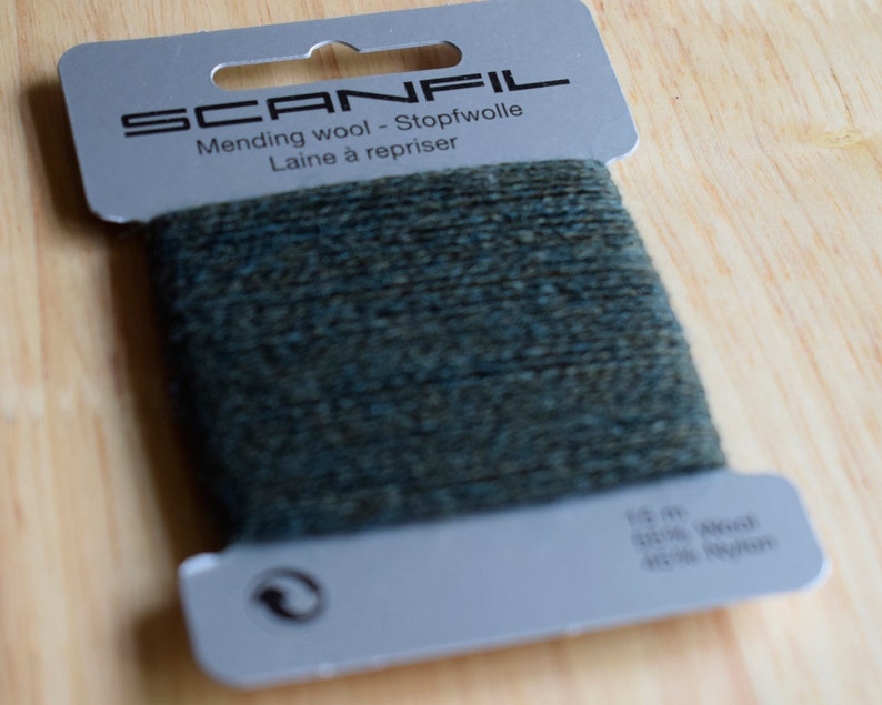 May include: A card of dark green mending wool by Scanfil. The grey cardboard packaging features the text SCANFIL, Mending wool - Stopfwolle, Laine &agrave; repriser, 15 m, 50% Wool, 50% Nylon, and a recycling symbol. The dark green yarn is wrapped around the centre.