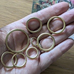 May include: A set of seven brass rings of varying sizes, displayed on a hand. The rings are smooth and shiny, with a warm golden hue.