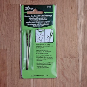 May include: Clover darning needles with latch hook eye in a clear plastic package. The package contains two silver needles with a hook design. Text on the package includes instructions and the Clover logo. Designed for threading multi-ply and bulky yarns.
