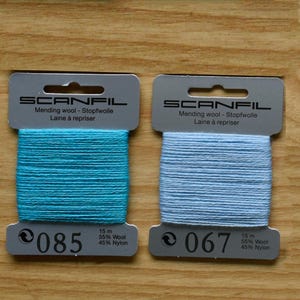 May include: Two Scanfil mending wool cards, one with turquoise thread labelled 085 and one with light blue thread labelled 067. Each card contains 15 metres of 55% wool and 45% nylon blend thread, featuring the brand name and product details printed on top.