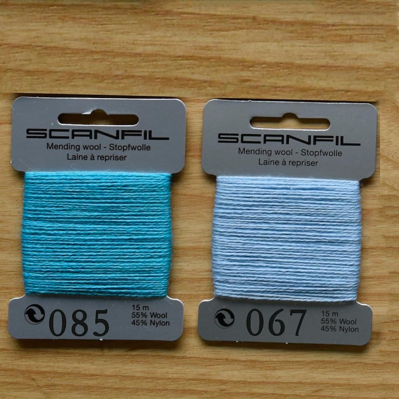 May include: Two Scanfil mending wool cards, one with turquoise thread labelled 085 and one with light blue thread labelled 067. Each card contains 15 metres of 55% wool and 45% nylon blend thread, featuring the brand name and product details printed on top.
