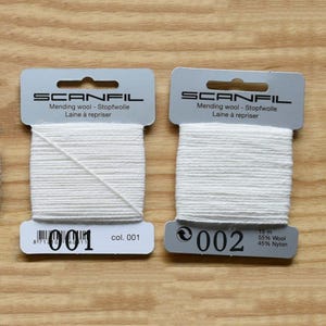 May include: Two rectangular cardboard cards wrapped with white mending wool. Each card features the Scanfil brand name, the text Mending wool - Stopfwolle - Laine &agrave; repriser, and colour numbers 001 and 002. The second card specifies 15 m, 55% Wool, 45% Nylon.