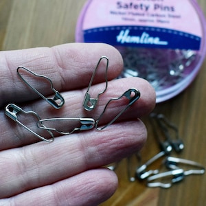 May include: A hand holding a handful of silver safety pins. The pins are in a variety of sizes and are all closed. The hand is holding the pins in front of a pink container with the text "Safety Pins" and "Hamline" on it.