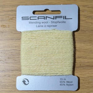 May include: A card of pale yellow mending wool by Scanfil. The light-coloured yarn is wrapped around a silver-toned cardboard holder featuring the brand name, 15 m length, and a composition of 55% wool and 45% nylon printed on the front surface.