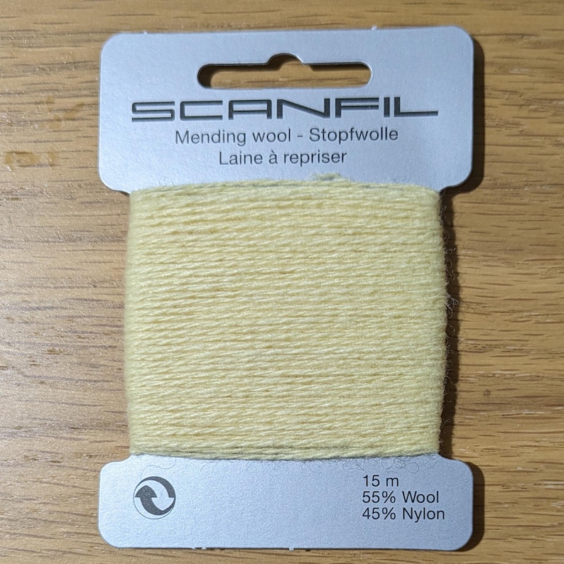 May include: A card of pale yellow mending wool by Scanfil. The light-coloured yarn is wrapped around a silver-toned cardboard holder featuring the brand name, 15 m length, and a composition of 55% wool and 45% nylon printed on the front surface.