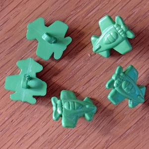 May include: Five green plastic buttons are displayed. Two are clover-shaped, and three are aeroplane-shaped. The buttons are arranged on a wooden surface.