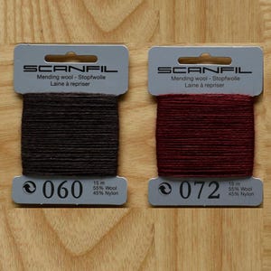 May include: Two Scanfil mending wool cards, one dark brown labelled 060 and one deep red labelled 072. Each card contains 15 metres of 55% wool and 45% nylon thread. The packaging displays the brand name, material composition, and colour code.
