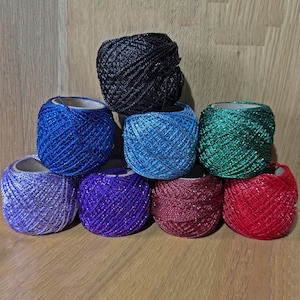 May include: A selection of eight spools of shimmering yarn in a variety of colours. The yarn shades include black, blue, green, purple, and red. The spools are arranged in a neat display, suitable for various craft projects.