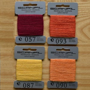 May include: Four Scanfil mending wool cards in burgundy, orange, yellow, and light orange. Each card contains 15 metres of 55% wool and 45% nylon thread. The cards are labelled with colour numbers 057, 093, 087, and 090, featuring a top hanging hole.