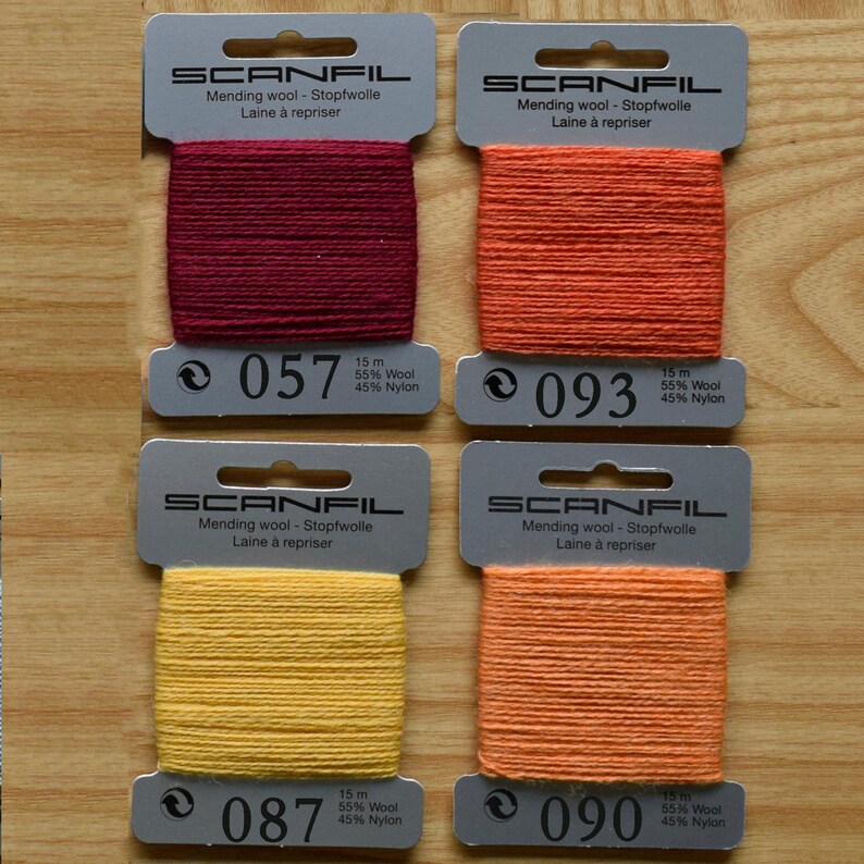 May include: Four Scanfil mending wool cards in burgundy, orange, yellow, and light orange. Each card contains 15 metres of 55% wool and 45% nylon thread. The cards are labelled with colour numbers 057, 093, 087, and 090, featuring a top hanging hole.