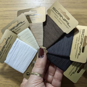 May include: A variety pack of six spools of cotton mending thread in different colours, including white, beige, brown, and grey. Each spool is labelled "SCANFIL Cotton mending - Baumwollstopfgarn Coton à repriser".