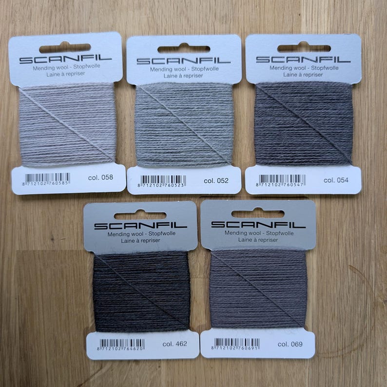 May include: Five rectangular cards of Scanfil mending wool in varying shades of grey. Each card features the brand name, the text Mending wool - Stopfwolle - Laine &agrave; repriser, a barcode, and a specific colour number: 058, 052, 054, 462, and 069.