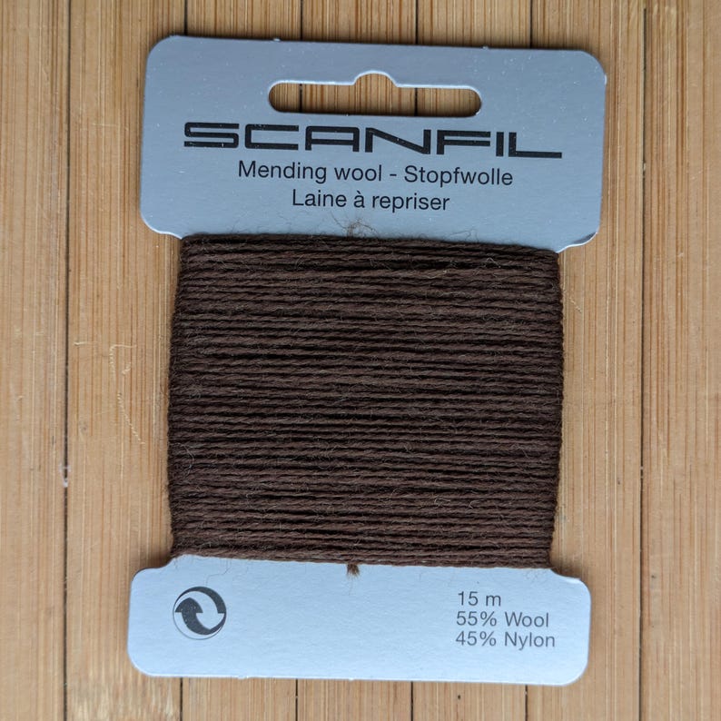 May include: A card of dark brown mending wool by Scanfil, featuring 15 m of thread composed of 55% wool and 45% nylon. The product is wound around a light grey rectangular cardboard holder with text printed in black, including the brand name.