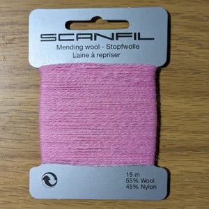May include: A card of Scanfil mending wool featuring pink yarn wrapped around a grey cardboard holder. The text reads Scanfil, Mending wool, Stopfwolle, Laine &agrave; repriser, 15 m, 55% Wool, and 45% Nylon. The card has a hole at the top for hanging.