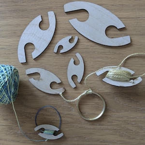 May include: A set of wooden yarn bowls with a gold metal ring. The bowls are in various sizes and have a slot for holding yarn. There are two balls of yarn, one blue and green and one beige.
