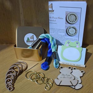 May include: A craft kit featuring a green frog, blue and green embroidery floss, a gold tin with a frog logo, a needle, brown rings, gold rings, and two wooden frog ornaments with the text "The Frog in the Craft".