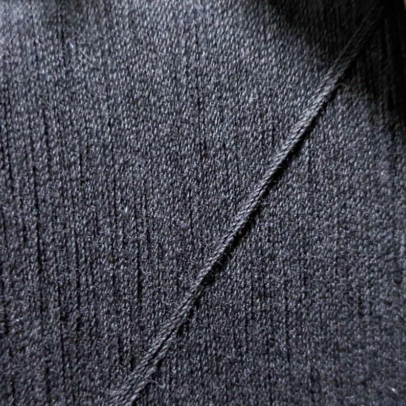 May include: Close-up of a black fabric with a diagonal seam. The fabric has a textured, woven appearance.