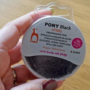 以下が含まれることがあります： PONY製の黒いスチール製ドレスメーカーピンが入った容器。容器には「PONY Black STEEL」と「create beauty with PONY」の文字が書かれている。容器は35グラムで、16本の0.65mmのピンが入っている。