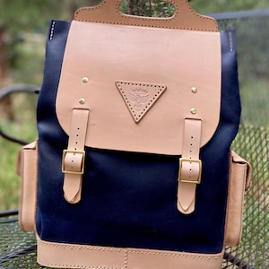 Könnte beinhalten: Ein dunkelblauer Canvas-Rucksack mit einer braunen Ledertasche und Besätzen. Der Rucksack hat zwei verstellbare Träger und einen Tragegriff oben. Die Klappe hat ein Dreieckslogo mit dem Text "The Frontier" darin.